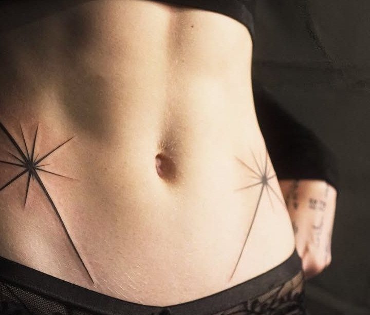 Two fine-line black ink starburst tattoos positioned on either side of a person's navel.