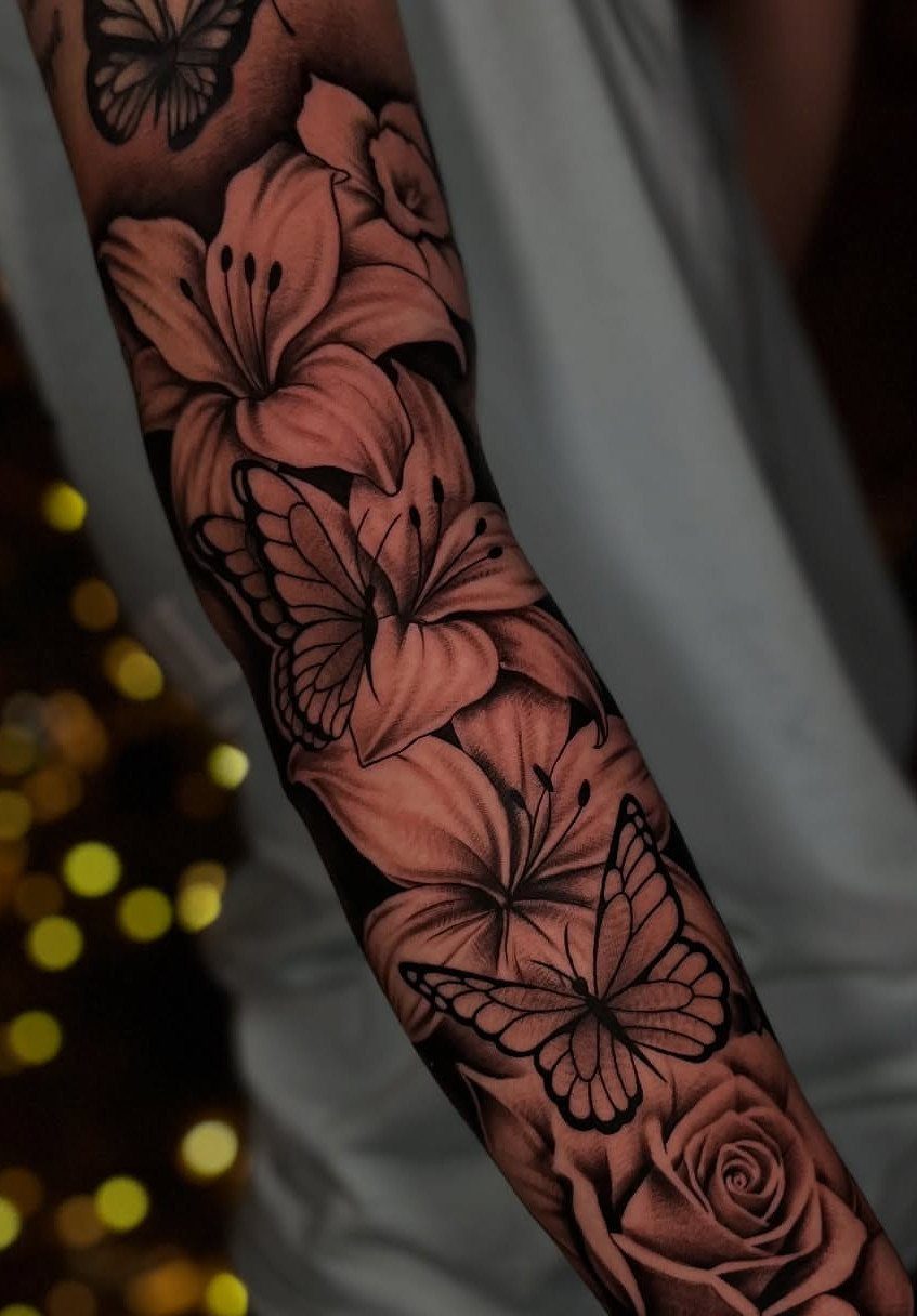 A detailed black and grey tattoo sleeve featuring lilies, roses, and butterflies on a person's arm.
