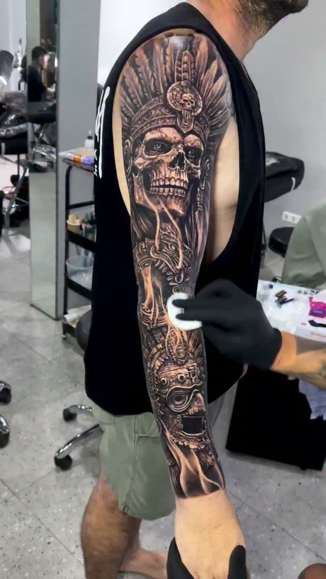 A man's arm displaying a detailed black and grey skull and headdress full sleeve tattoo.