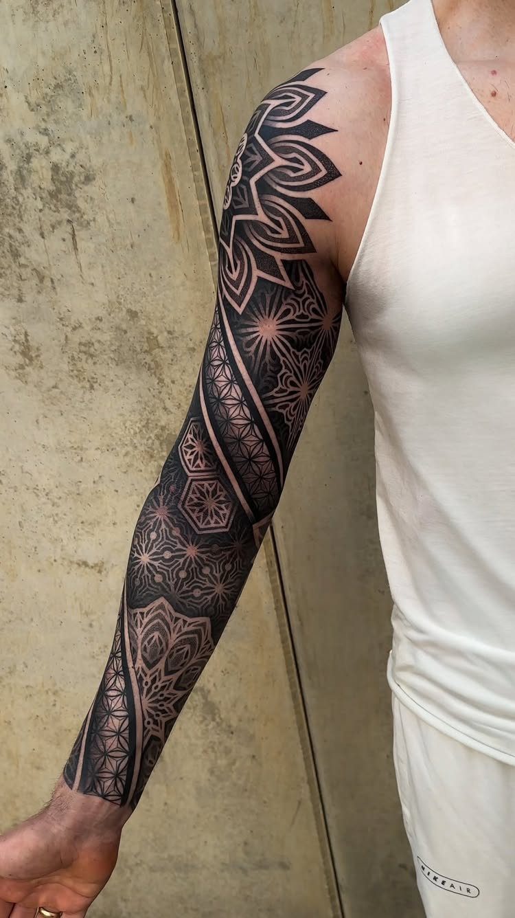 A person's arm displaying a detailed black and grey geometric sleeve tattoo with mandala patterns.