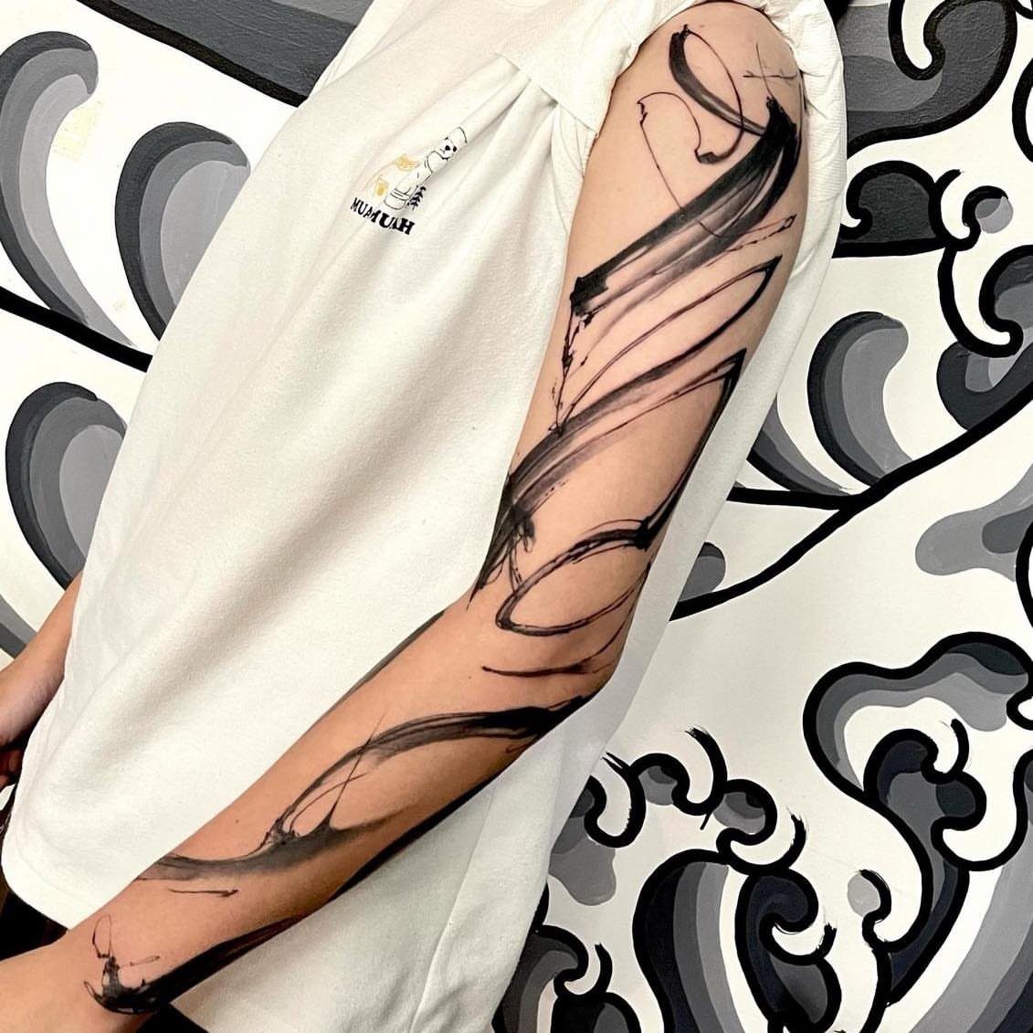 A person's arm displaying an abstract black ink brushstroke sleeve tattoo against a patterned background.