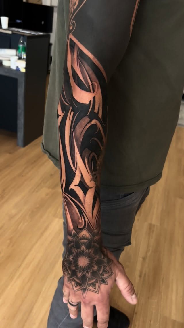 A person's arm displaying a large black and grey ornamental tattoo sleeve with a mandala on the hand.