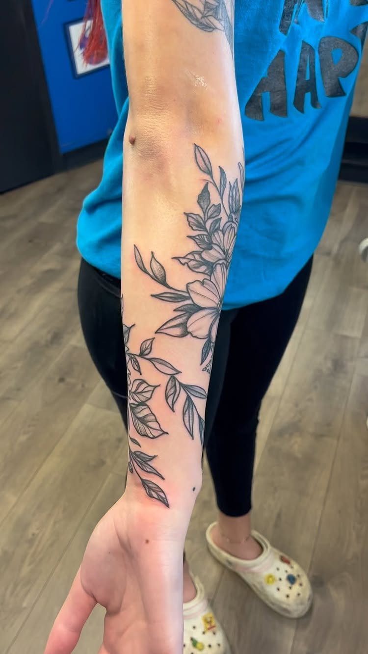 22 Tattoo Pattern Floral Ideas for Arms, Ribs & Full Sleeves 10 Black ink floral tattoo with leaves and blossoms wrapping around a person's forearm.