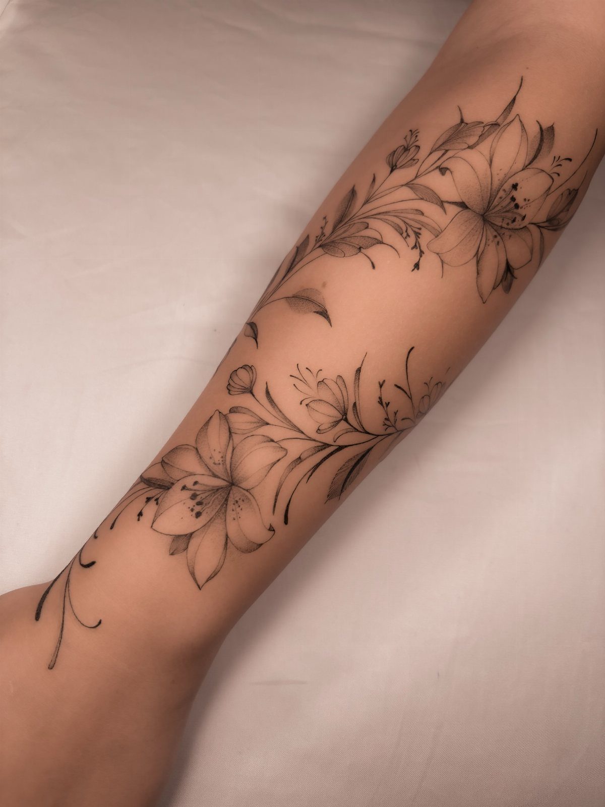 22 Tattoo Pattern Floral Ideas for Arms, Ribs & Full Sleeves 5 Black ink lily flower tattoos on a person's forearm with delicate leaves and stems.