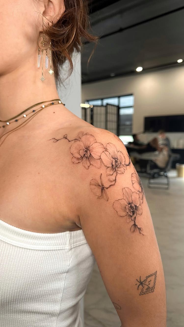 22 Tattoo Pattern Floral Ideas for Arms, Ribs & Full Sleeves 13 A woman's shoulder featuring a delicate black ink orchid flower tattoo design.