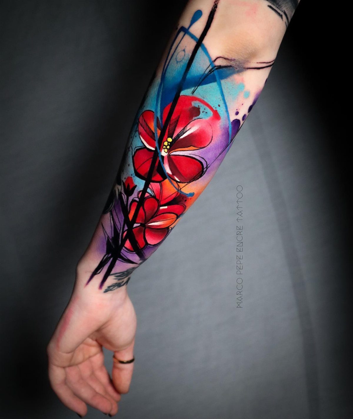22 Tattoo Pattern Floral Ideas for Arms, Ribs & Full Sleeves 6 A colorful watercolor floral tattoo with red flowers and blue splashes on a person's forearm.