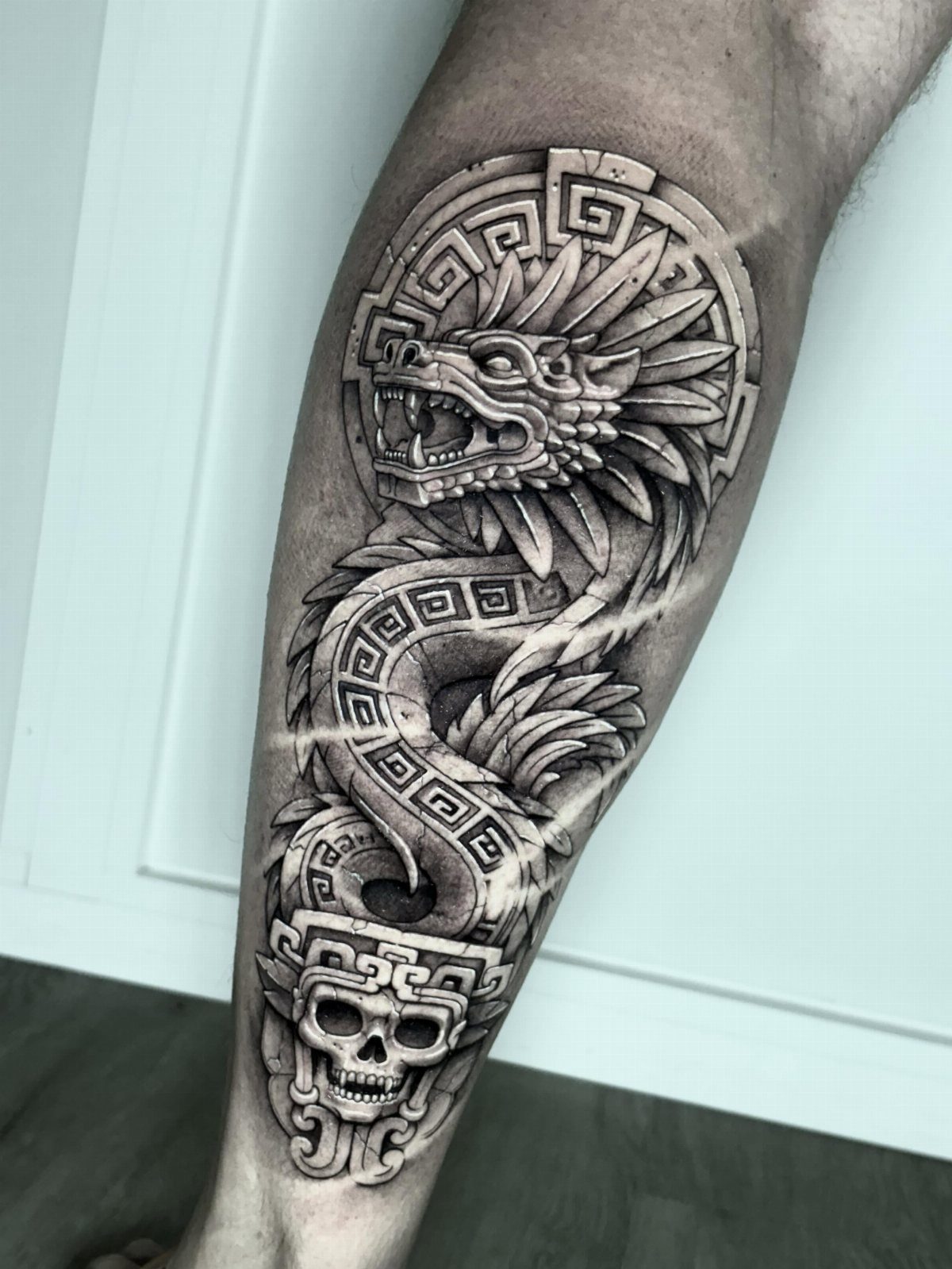 A large black and grey Aztec-style tattoo of a feathered serpent and skull on a leg.