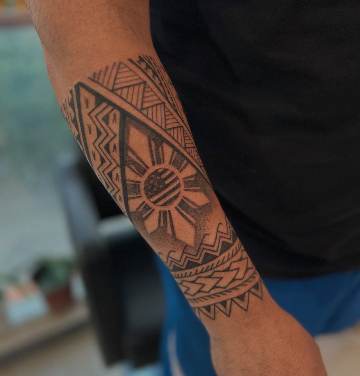 A black ink forearm tattoo featuring geometric patterns and a sun motif on a person's arm.