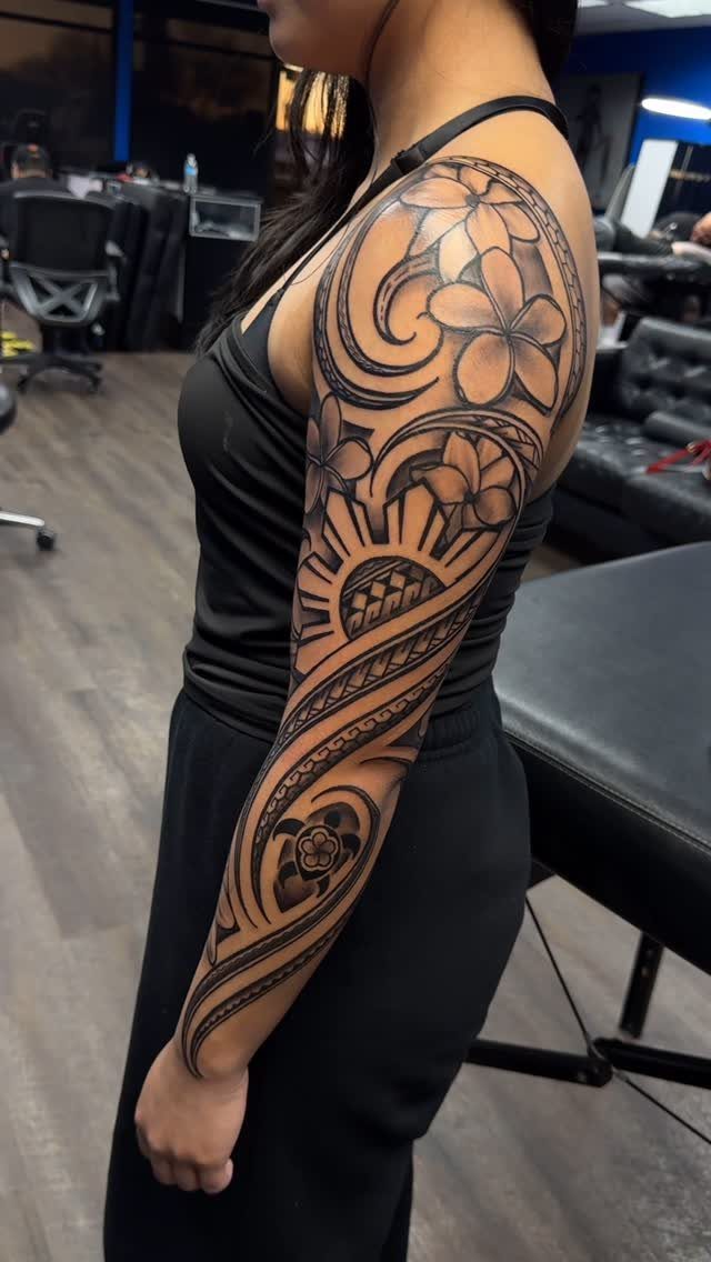 A woman's arm displaying a large black ink tribal sleeve tattoo with swirling and floral patterns.