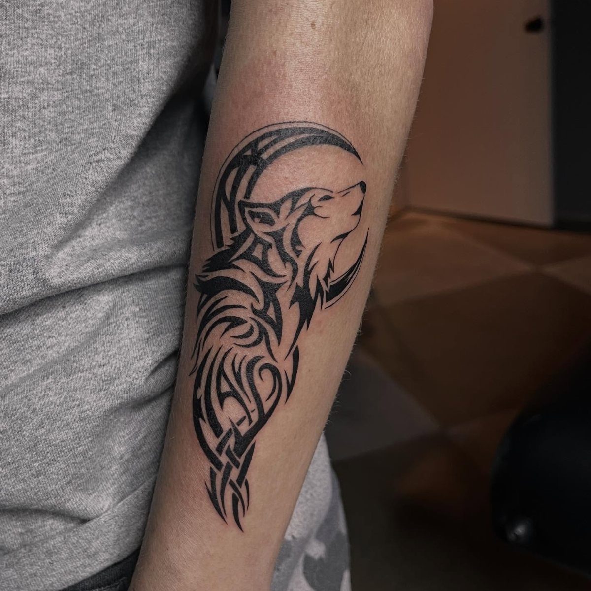 A black ink tribal tattoo of a wolf head and crescent moon on a forearm.