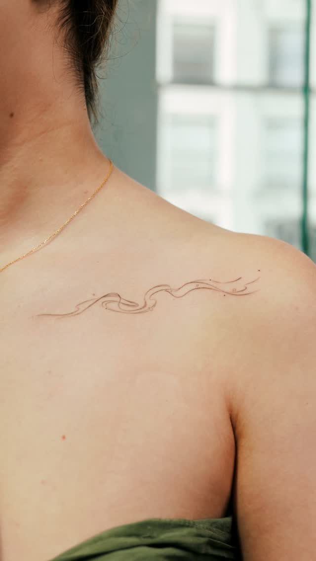 A fine-line wavy tattoo placed along a woman's collarbone with a gold necklace.