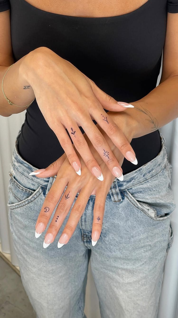 Hands with long French tip nails displaying small, delicate black ink finger tattoos.