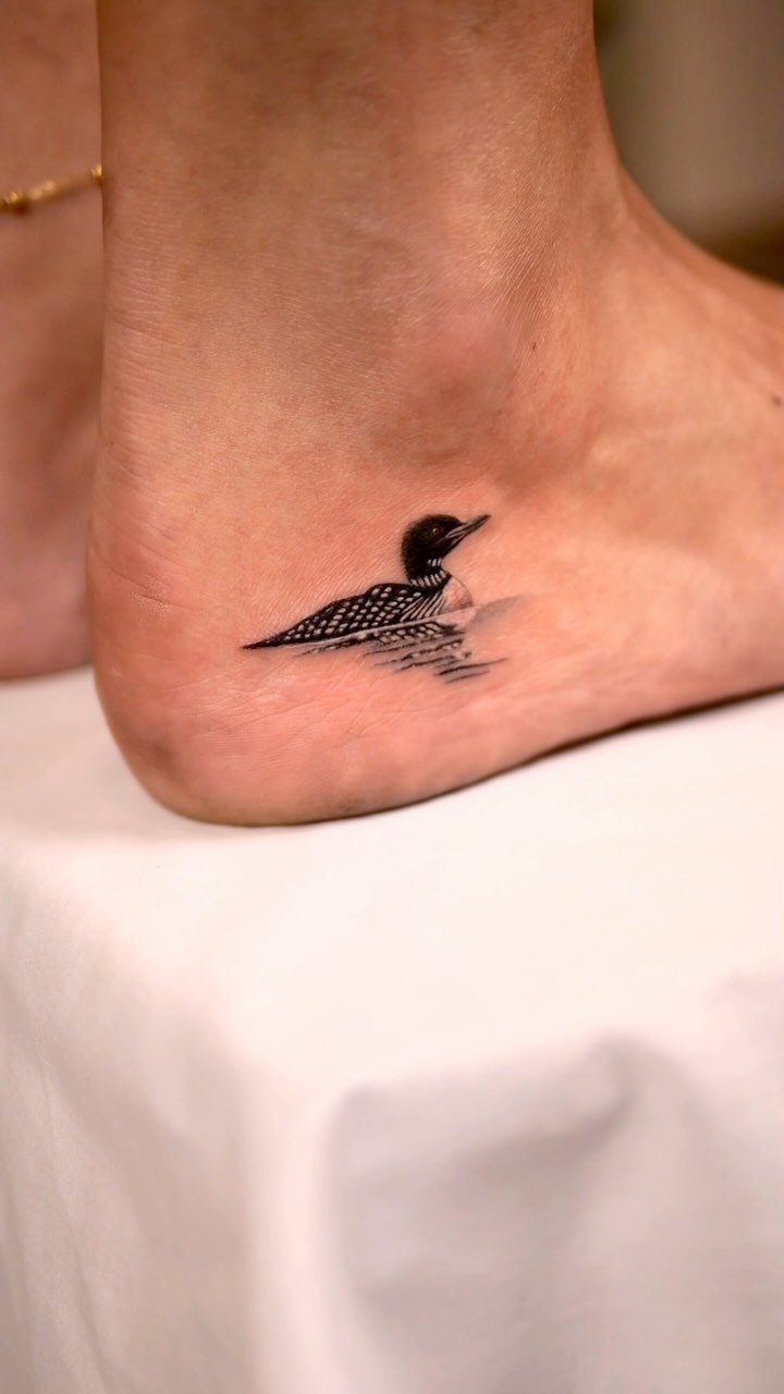 A small black and white loon tattoo on the side of a person's foot.