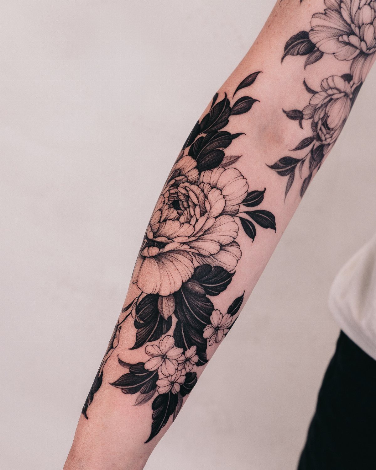 Large black ink floral tattoos featuring peonies and leaves on a person's forearm.