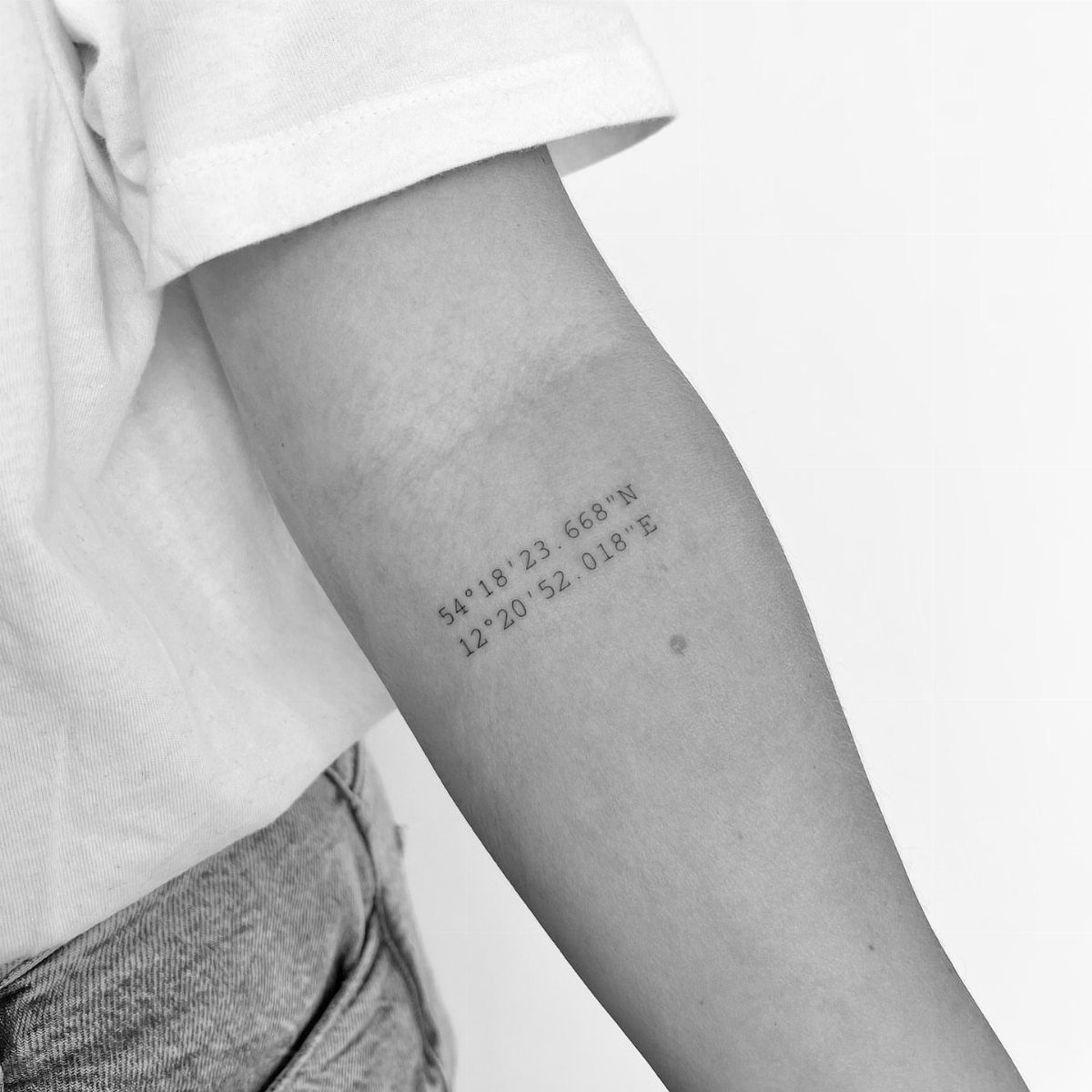 A person's inner forearm displaying a fine line tattoo of geographic GPS coordinates.