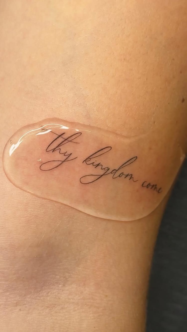 A fine line cursive tattoo reading 'thy kingdom come' on a person's skin under a clear film.