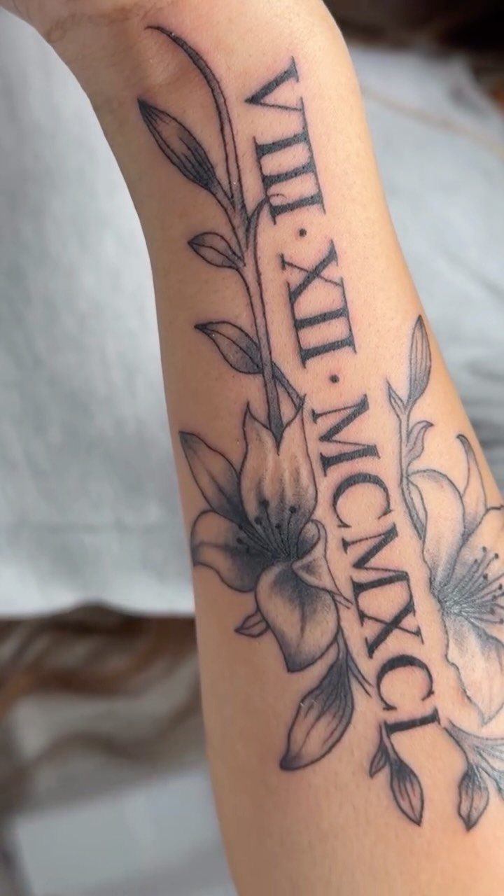 Roman numeral tattoo with lily flowers on a person's inner forearm.