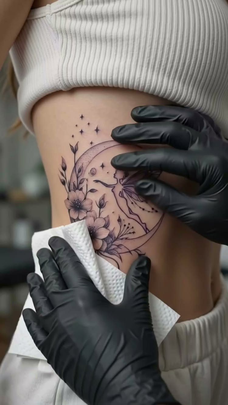 A fairy silhouette tattoo with a crescent moon and flowers on a person's ribs.