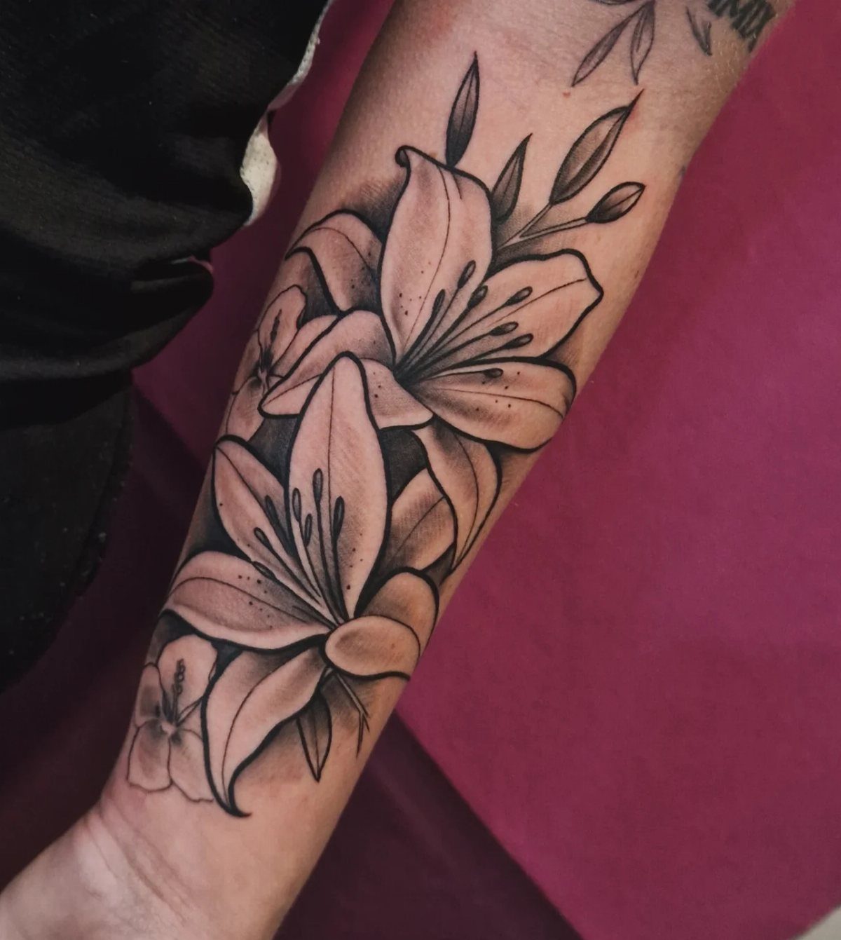 Black and grey lily flower tattoo with leaves and stems on a person's forearm.