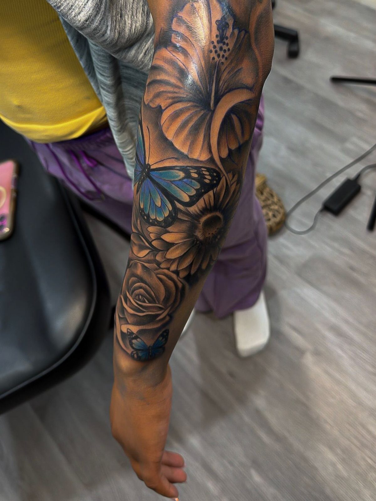 A detailed forearm tattoo featuring colorful hibiscus, roses, and blue butterflies on a person's arm.