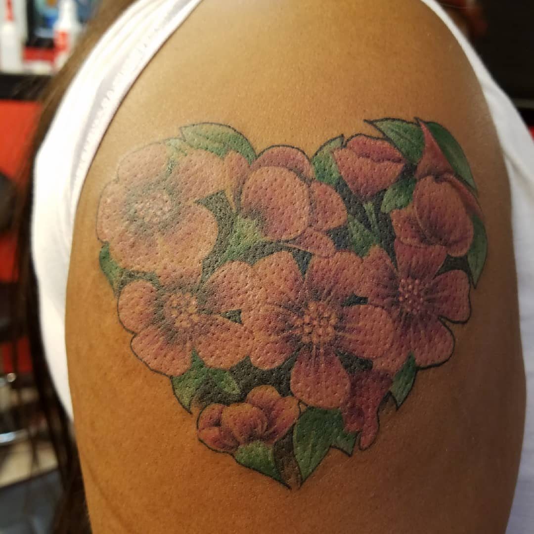 A heart-shaped tattoo of pink flowers and green leaves on a person's shoulder.