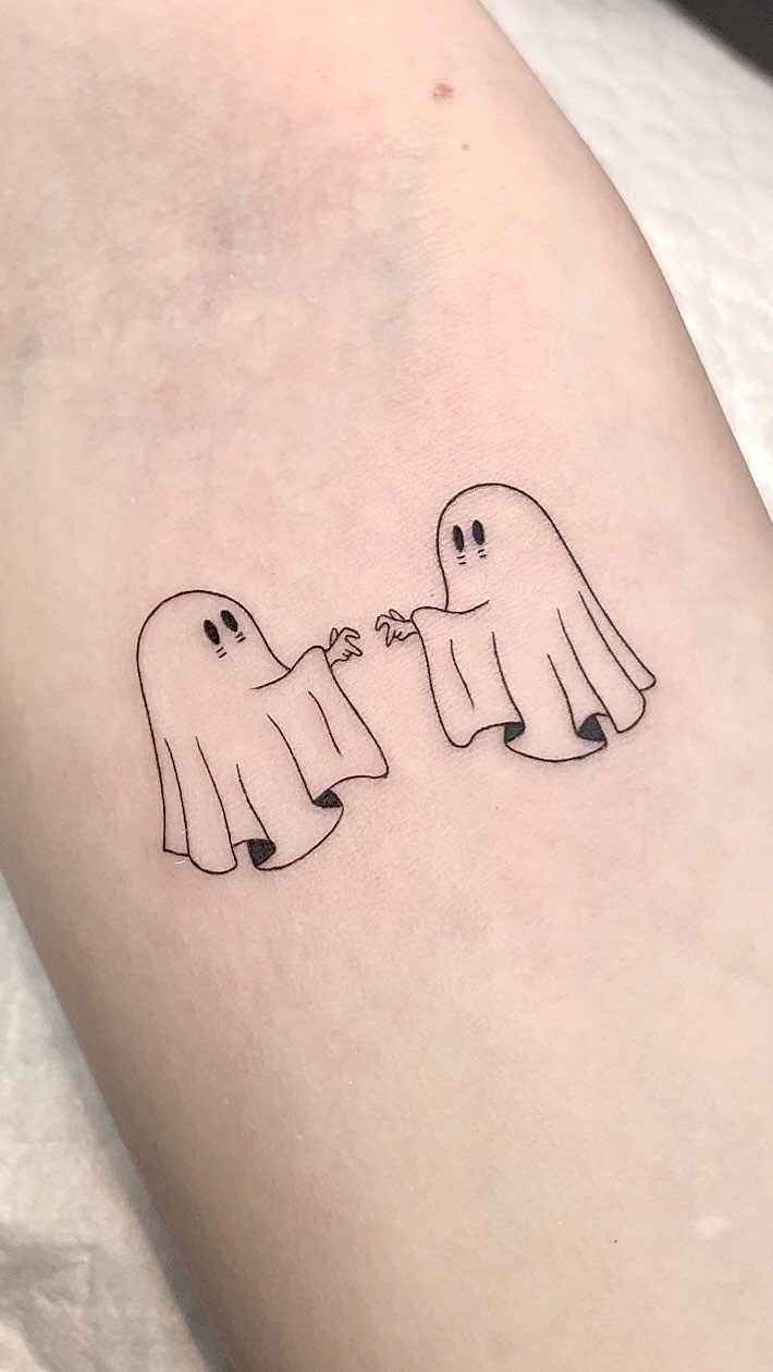 Two small minimalist black ink tattoos of cute friendly ghosts on skin.