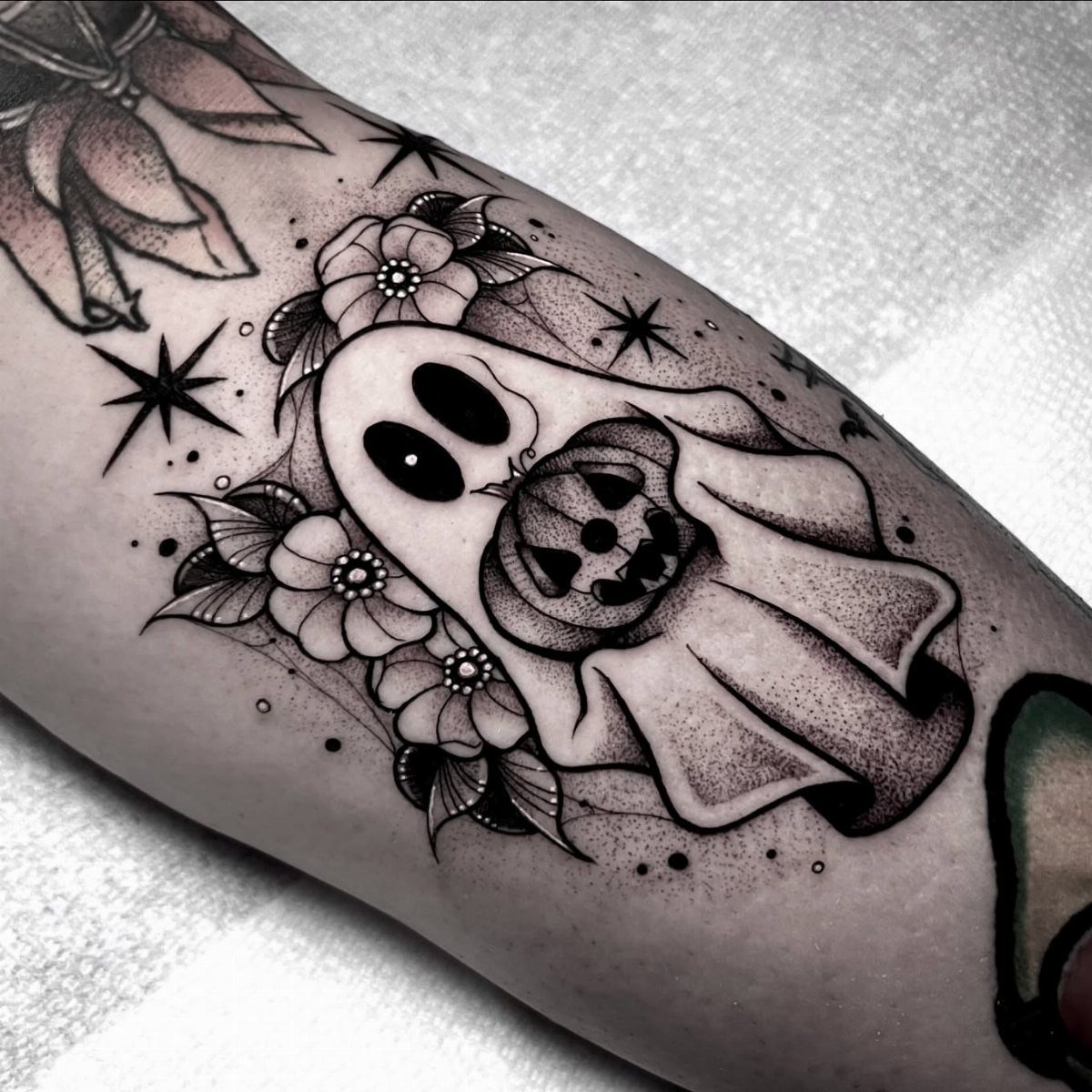 A black and grey tattoo of a cute ghost with flowers and stars on skin.
