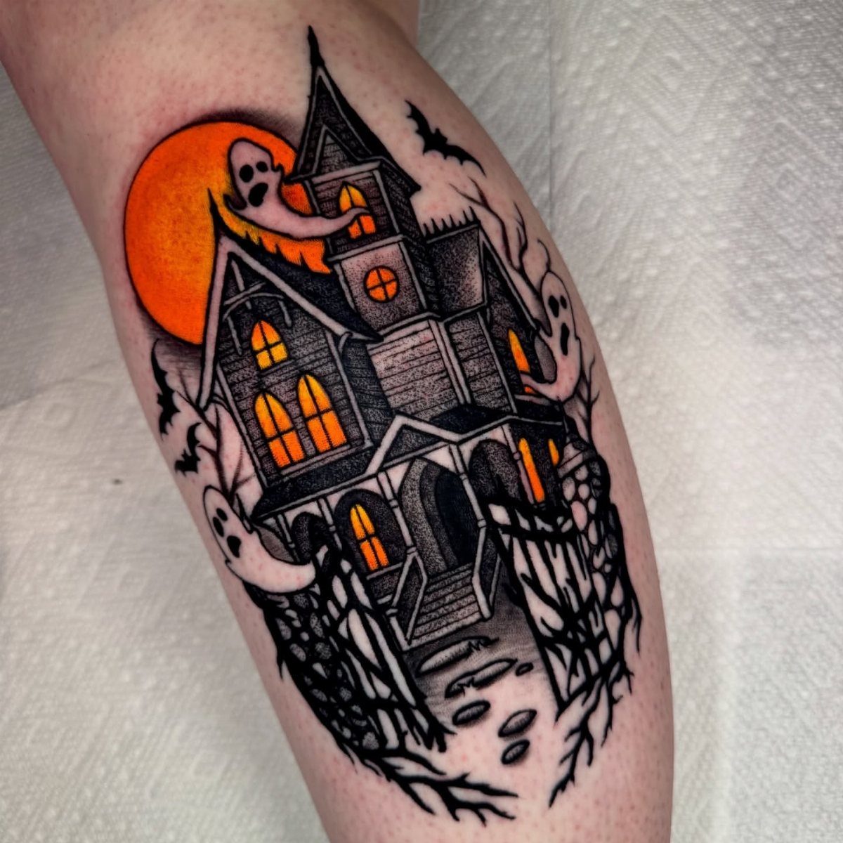 A detailed haunted house tattoo with glowing orange windows and ghosts on a person's leg.
