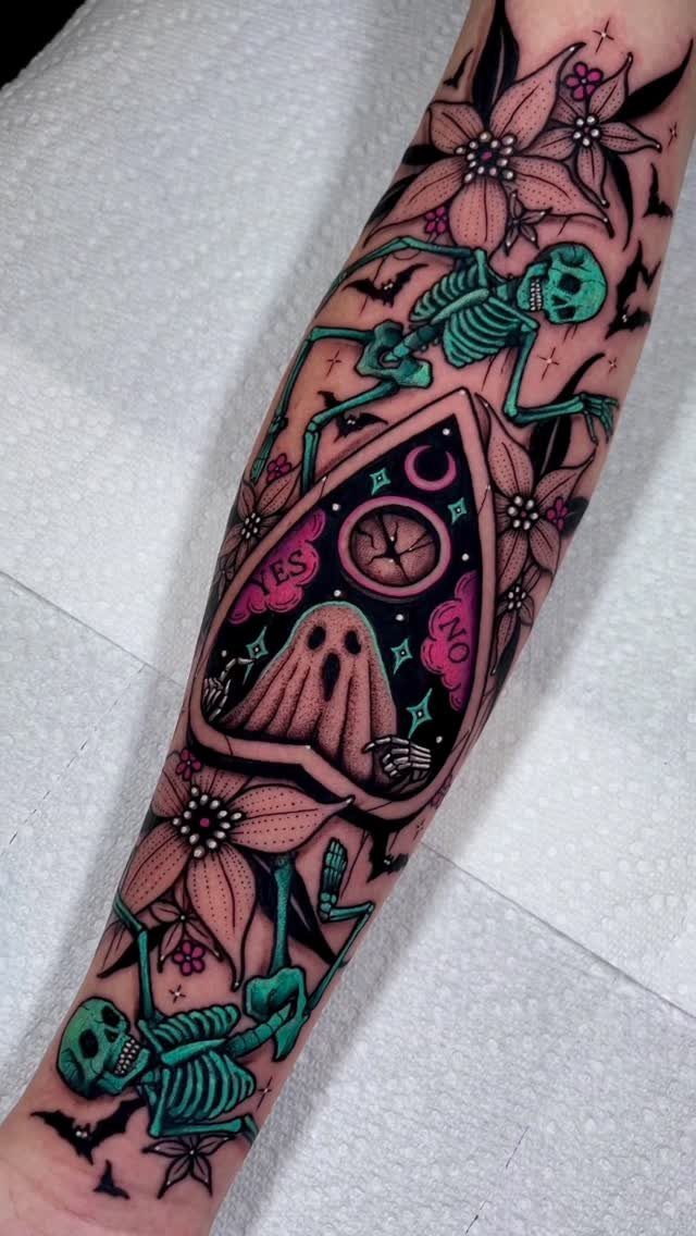 Colorful arm tattoo featuring small skeletons, a ghost in a triangle, and pink flowers.