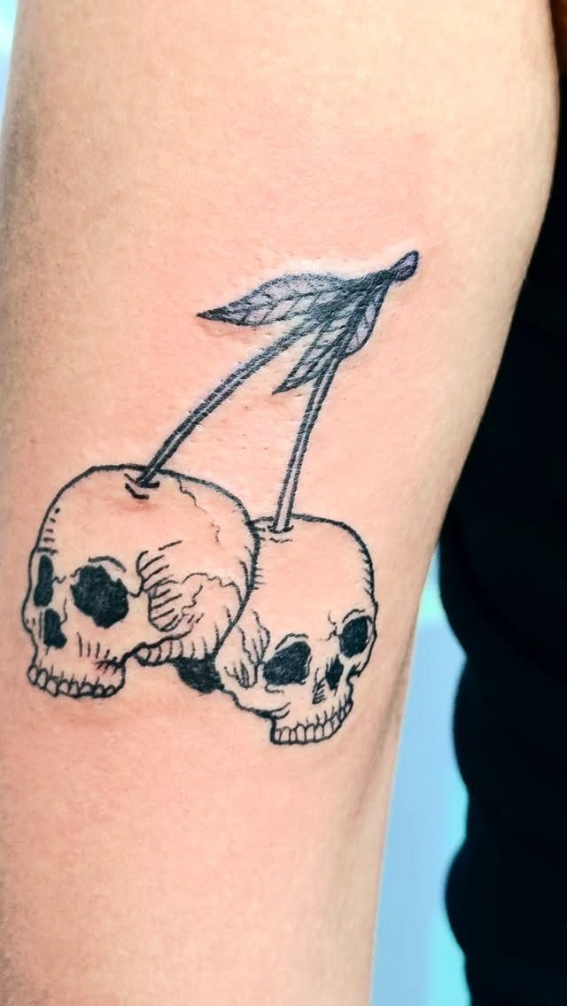 A black ink tattoo of two skull-shaped cherries on a person's arm.