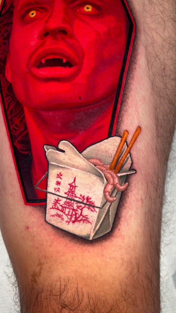 A vibrant tattoo of a red vampire face in a coffin above a takeout noodle box.