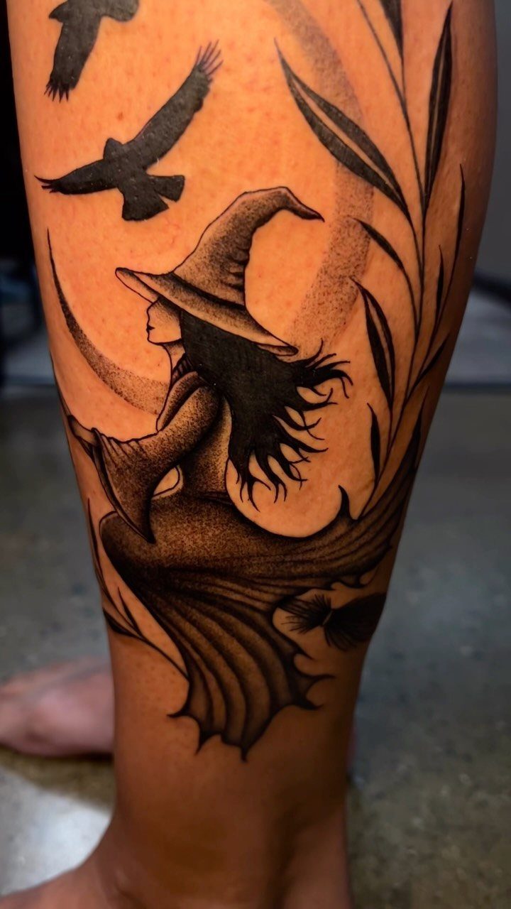 A black and grey silhouette tattoo of a witch and birds on a person's leg.