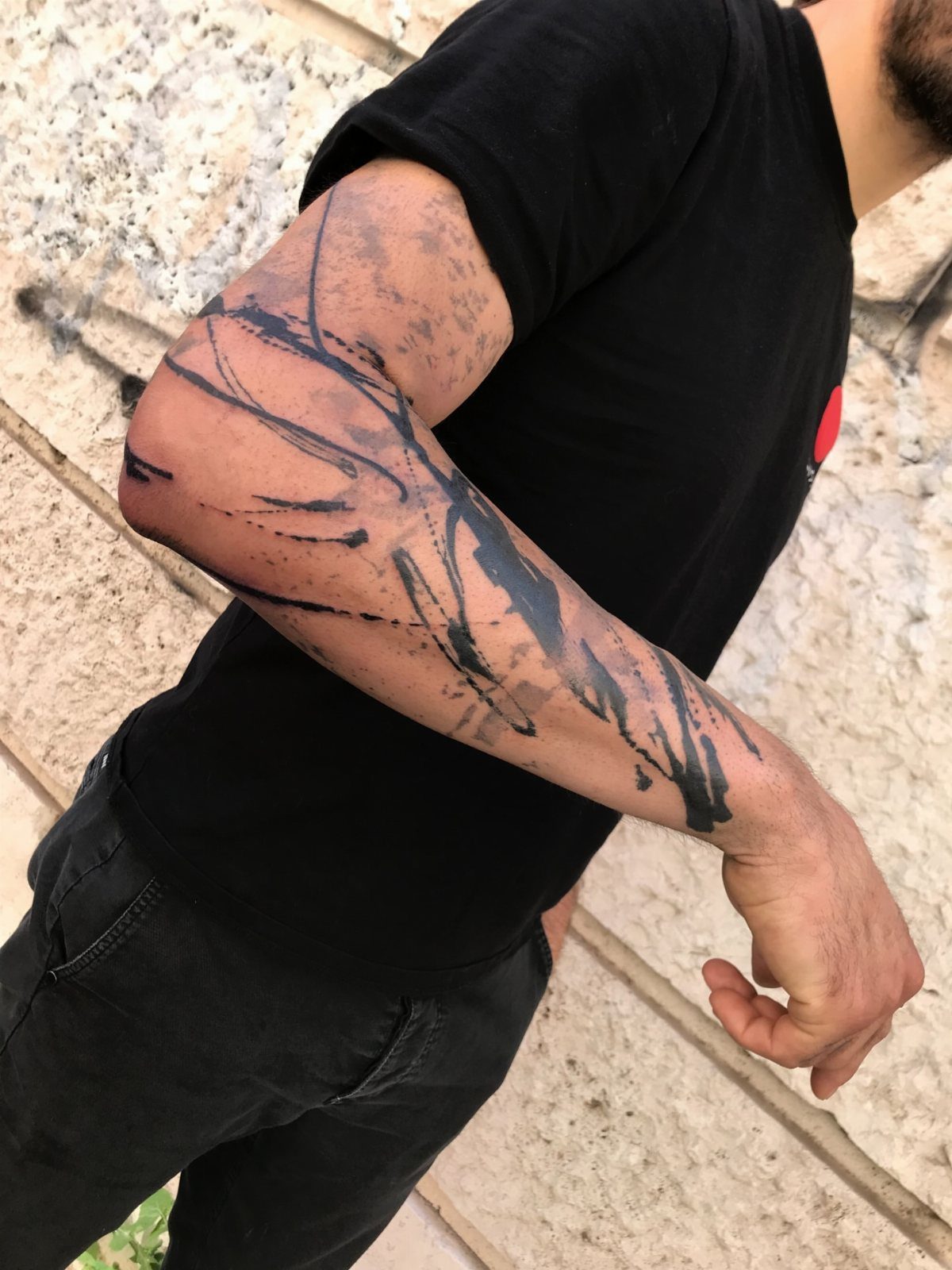 A man's arm featuring a large black abstract tattoo with brushstroke and splatter patterns.