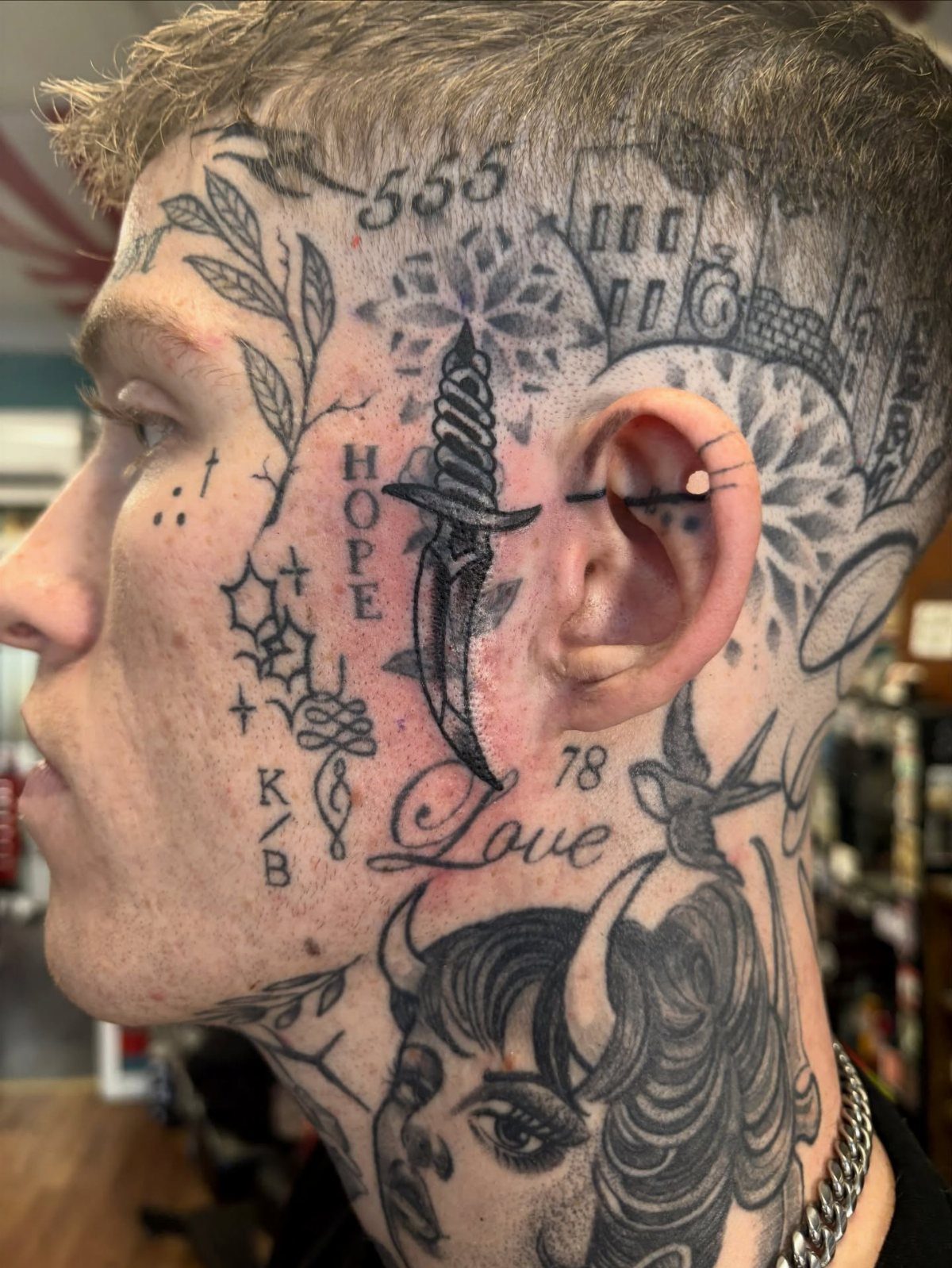 A man's profile showing various black ink tattoos including a dagger, floral patterns, and text.