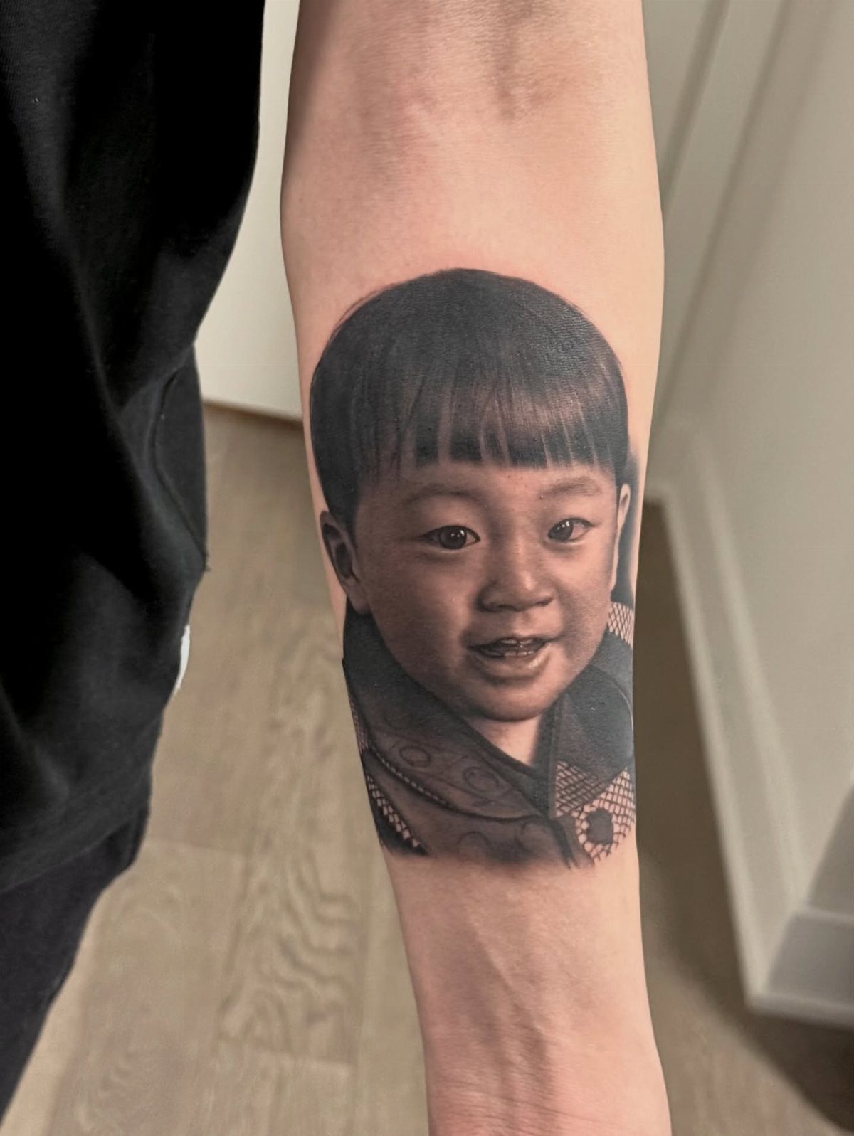 A realistic black and grey portrait tattoo of a smiling young child on a forearm.