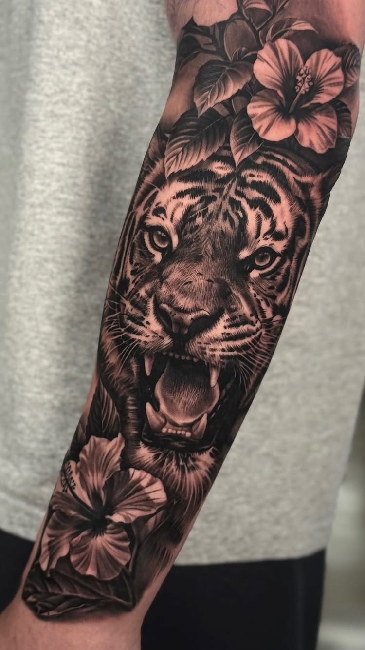 A realistic black and grey tiger tattoo with hibiscus flowers on a person's forearm.