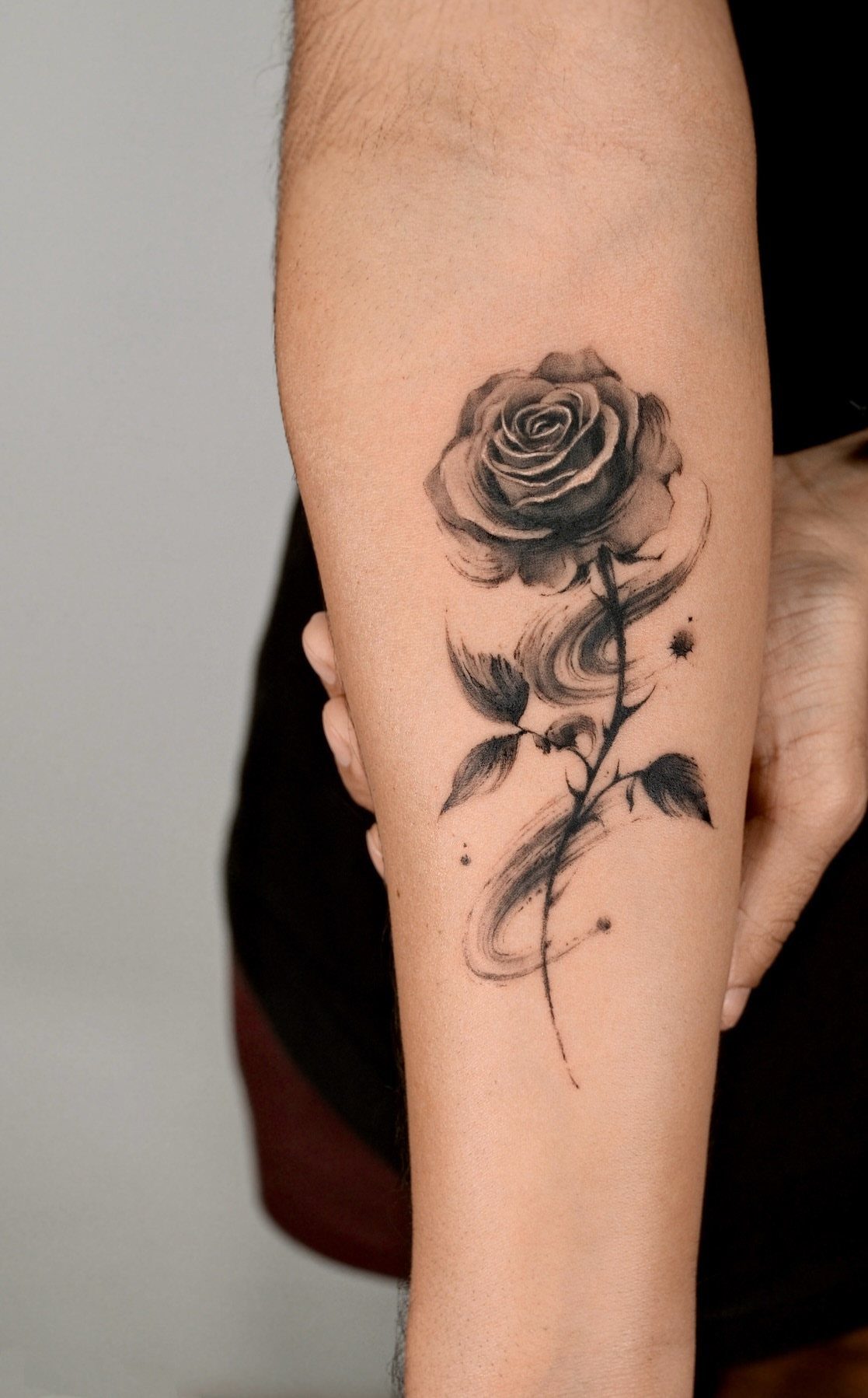 A black and grey rose tattoo with artistic flowing swirls on a person's forearm.