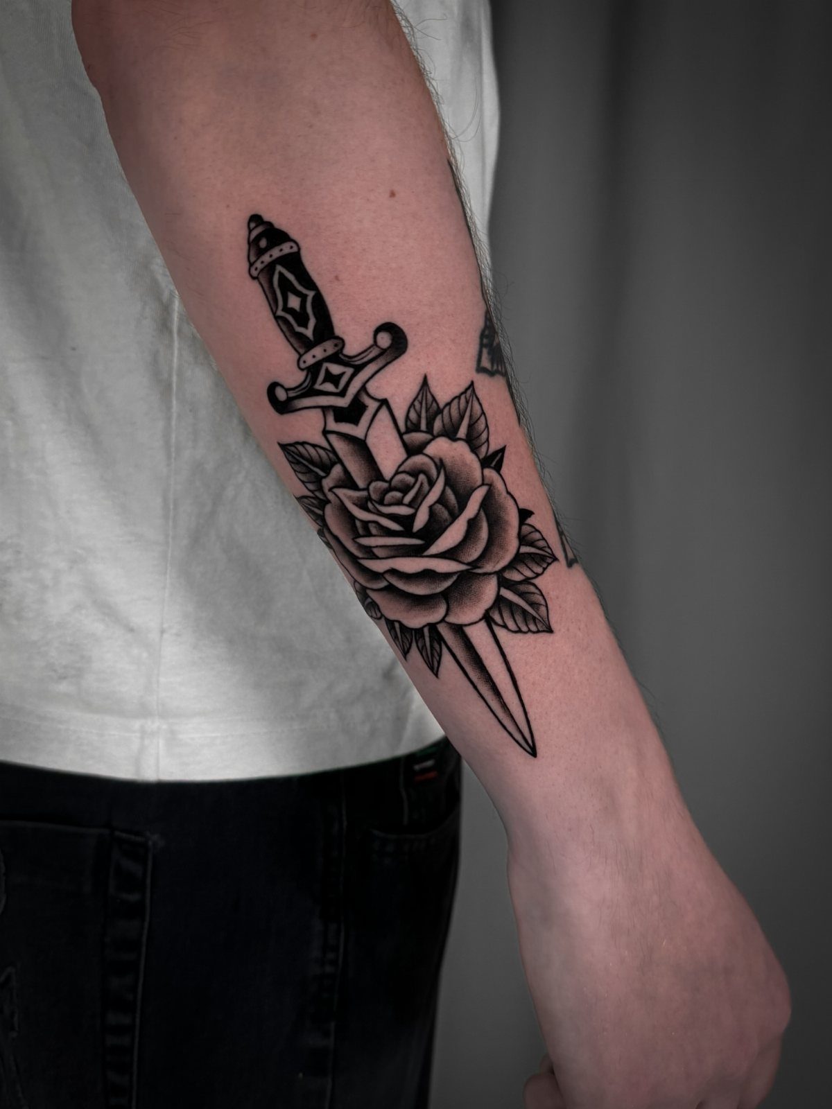 A black and grey tattoo of a rose and a dagger on a person's forearm.