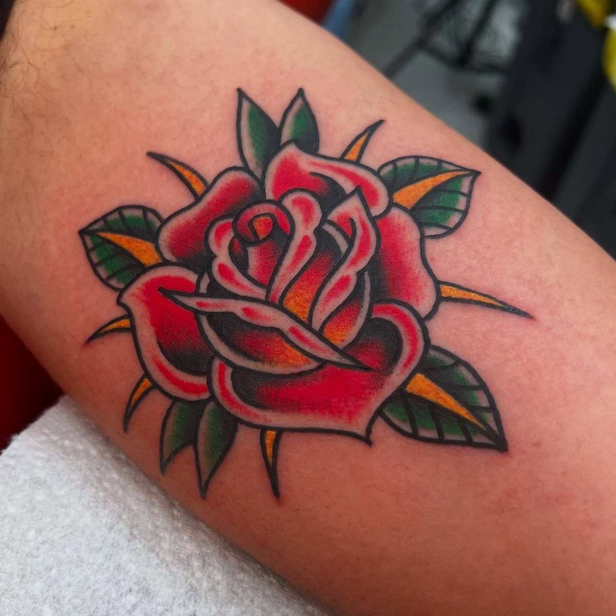 A traditional red rose tattoo with green leaves on a person's arm.