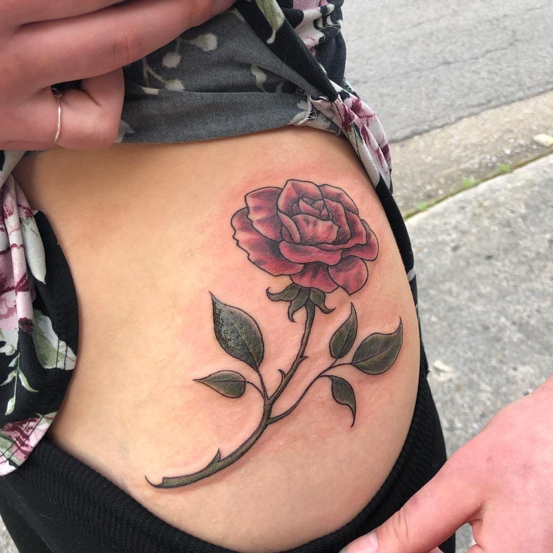 A pink rose tattoo with green leaves and a stem on a person's hip.