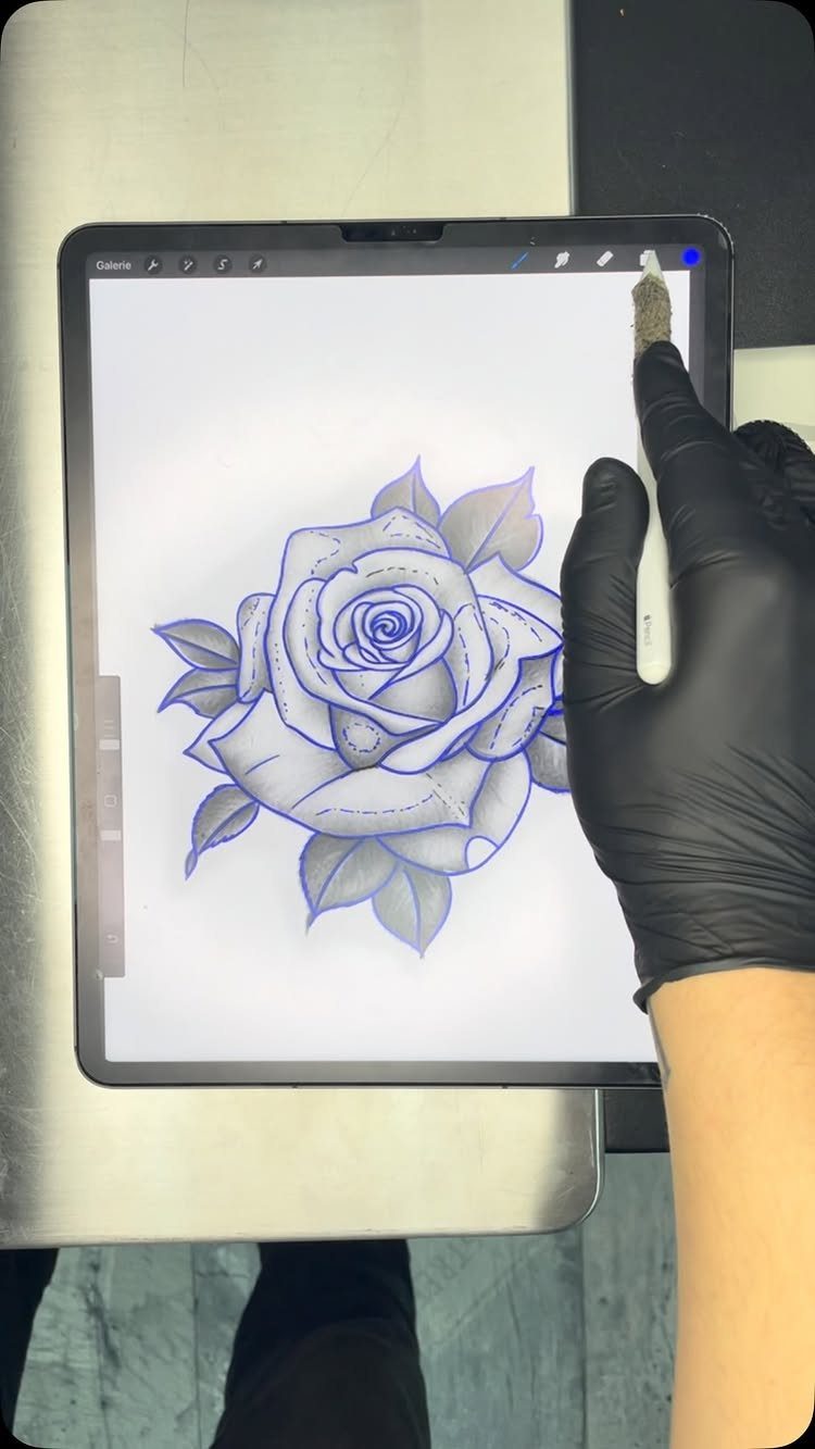 A gloved hand uses a stylus to draw a blue rose tattoo stencil on a digital tablet.