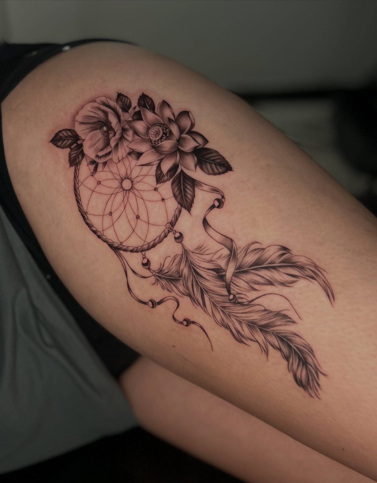 A black and grey dreamcatcher tattoo with flowers and feathers on a person's thigh.