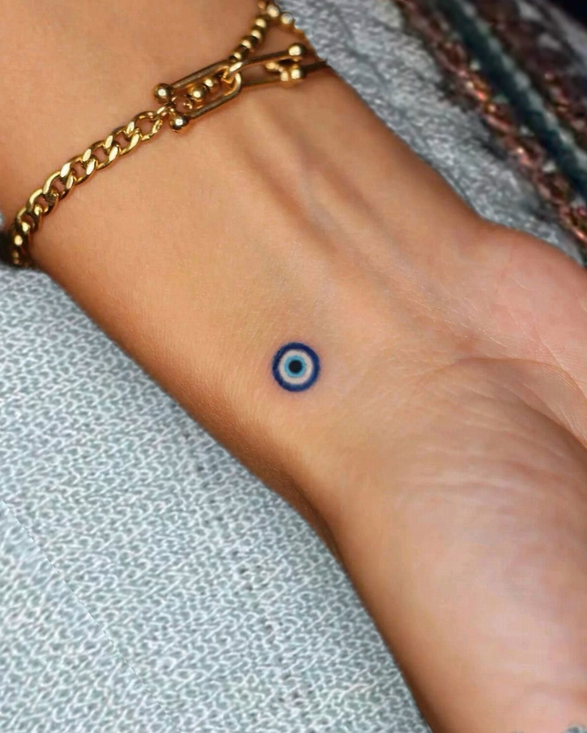 A small blue evil eye tattoo on a person's wrist with a gold bracelet.