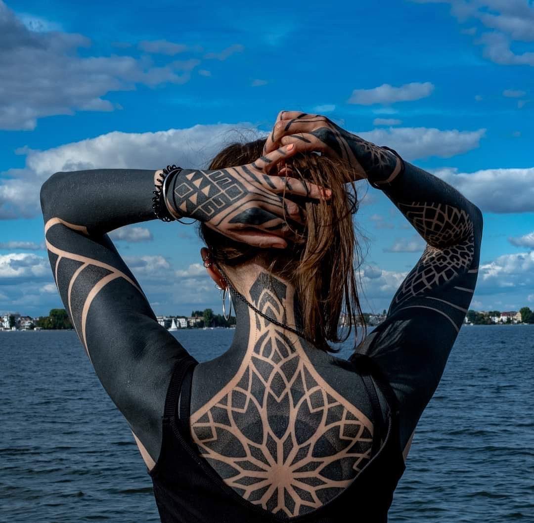 Woman with large blackwork geometric tattoos on her arms and back by a lake.