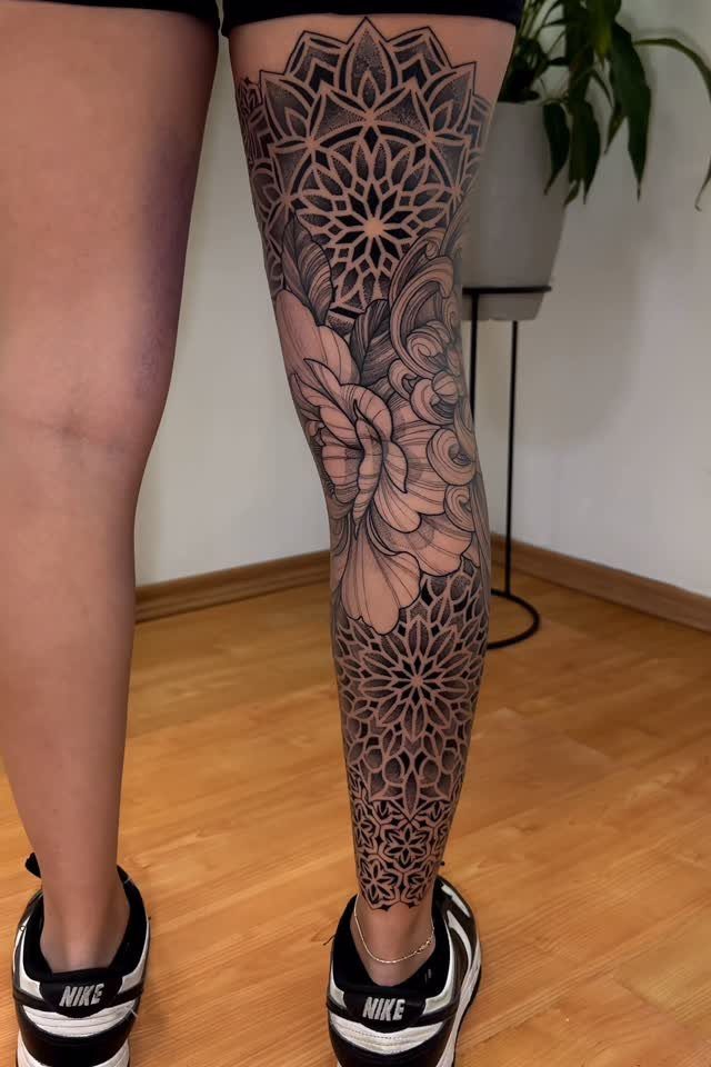 A person's leg decorated with a large, detailed black ink mandala and floral tattoo design.
