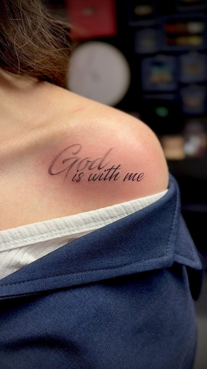 A script tattoo on a person's shoulder reading God is with me in black ink.