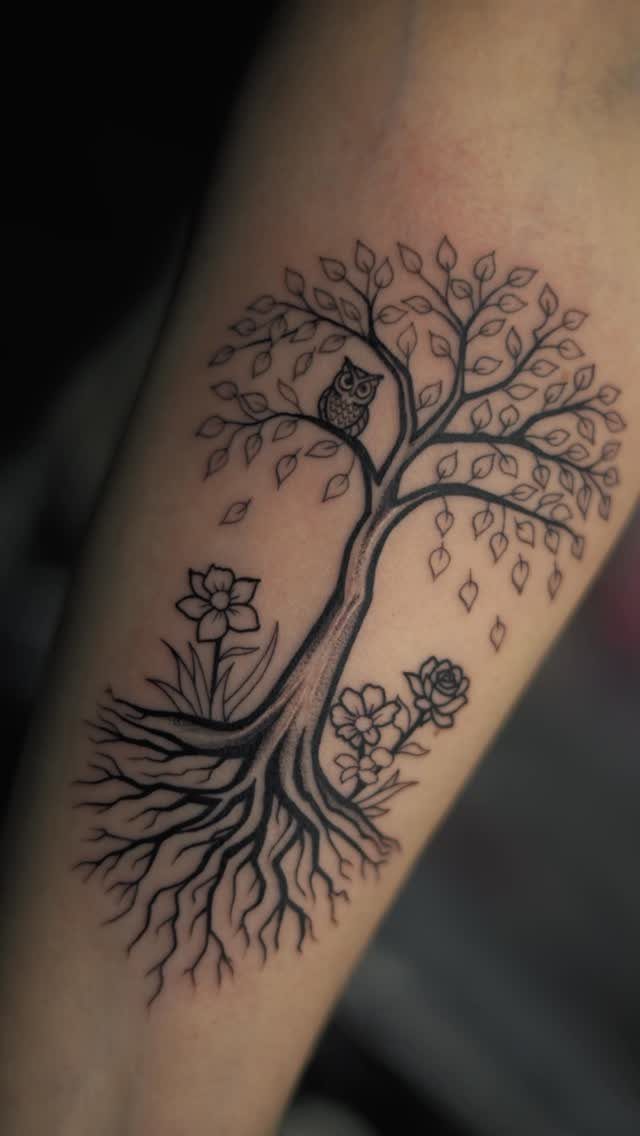 A black ink tree tattoo with roots, leaves, an owl, and flowers on a person's arm.