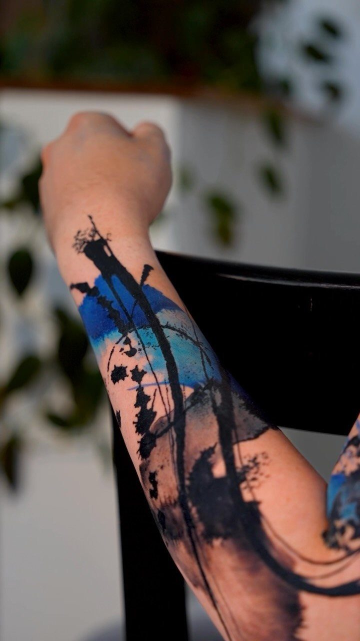 A person's forearm displaying a large, artistic watercolor style tattoo with blue and black ink splashes.