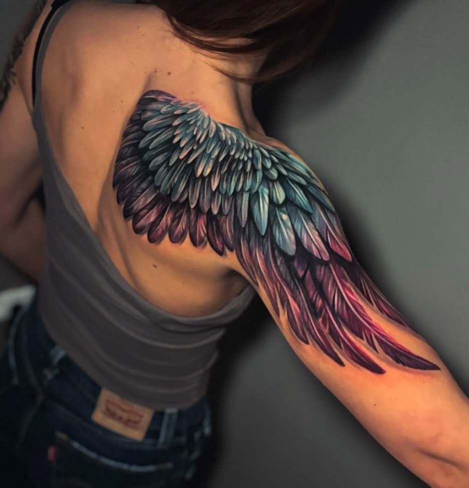A large, colorful feather wing tattoo covering a woman's shoulder and upper arm.