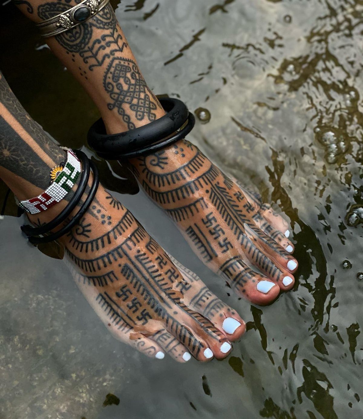 Tattooed feet and legs with geometric patterns submerged in rippling water.
